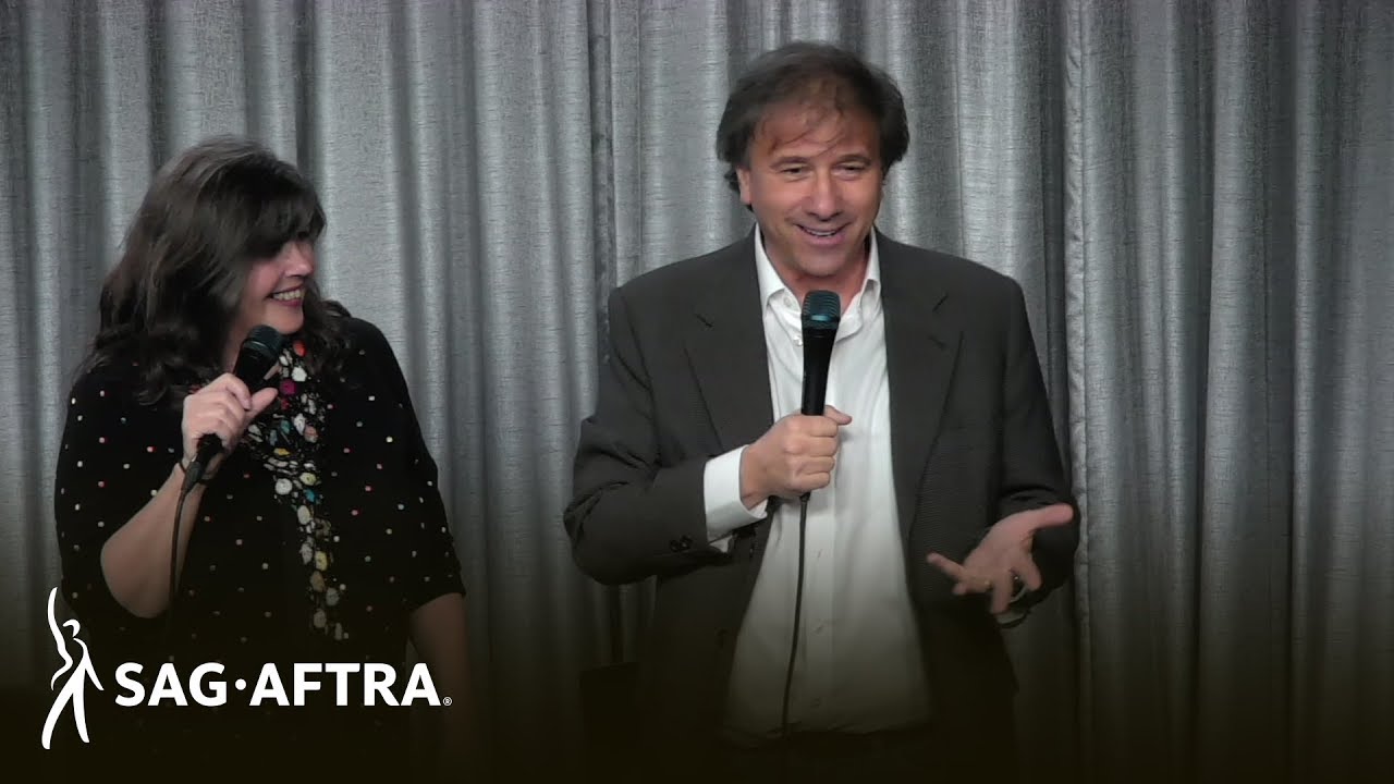 Megan a la izquierda sosteniendo el micrófono en la mano derecha mirando a Chuck a la derecha con un botón blanco y una chaqueta gris hablando a la audiencia