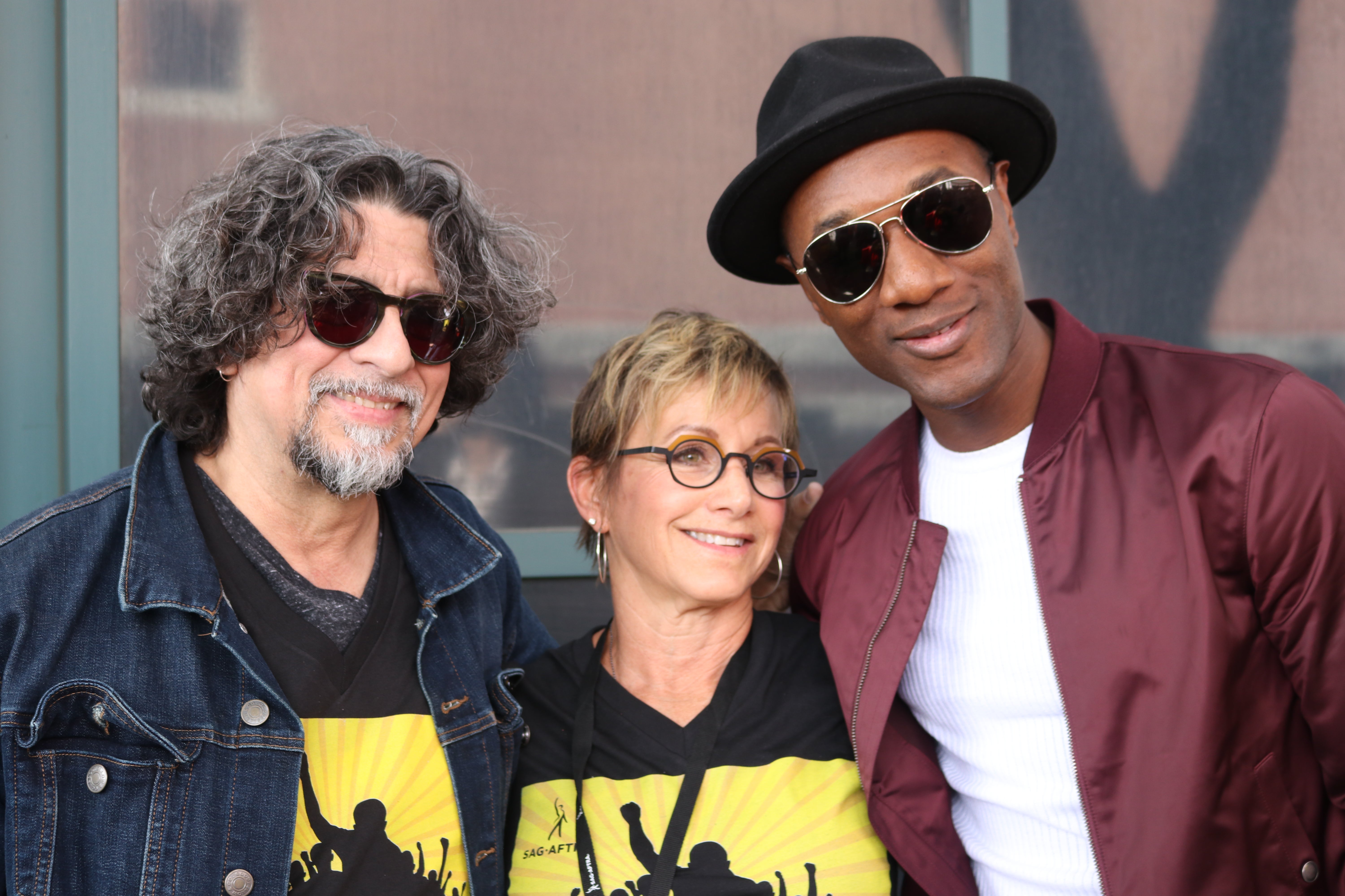 Navarro con una camiseta negra #SOSSBS y una chaqueta de jean sonriendo con gafas de sol oscuras. Carteris en el medio con una camiseta negra #SOSSBS y Blacc con una camisa blanca y una chaqueta marrón, tonos oscuros y un sombrero negro sonriendo de pie a la izquierda de Carteris