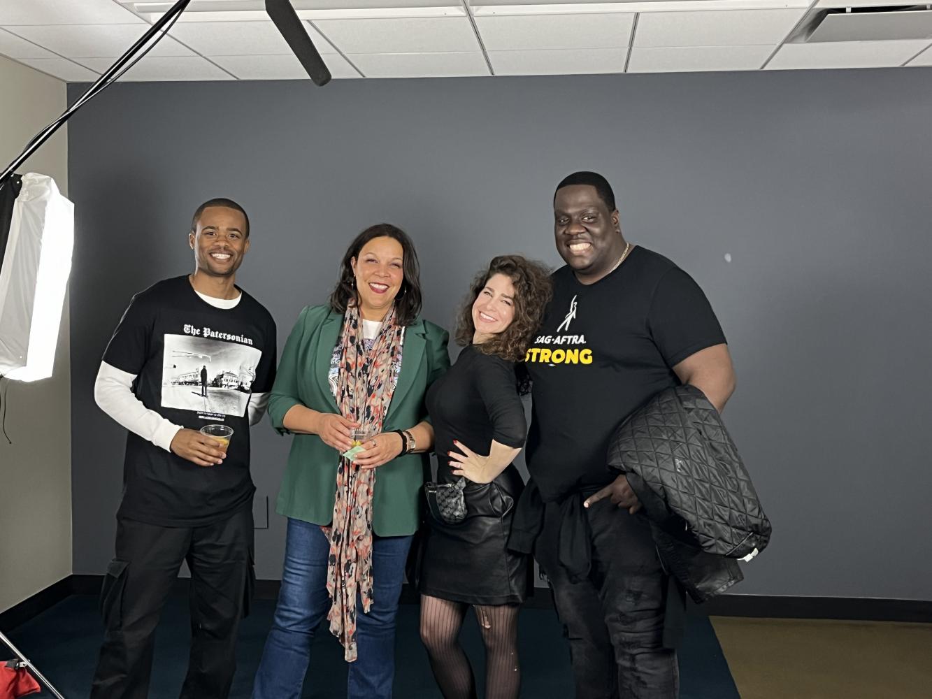 Four individuals, two African American men, Powell (Black) and Seeds stand in a large room. Powell, center left, wears a green top, colorful scarf and jeans; Seeds wears an all-black outfit. Seeds poses with her hand on her hip.