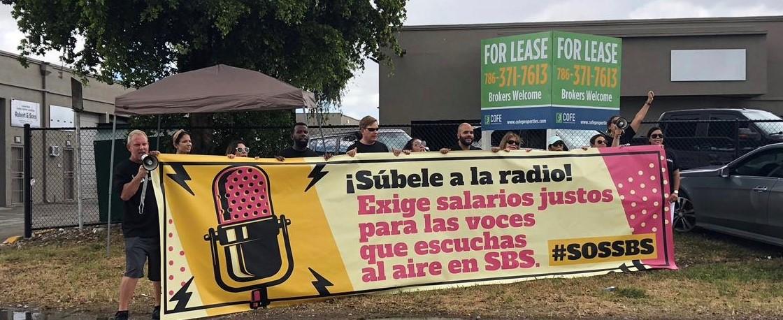 Miembros de SAG-AFTRA sosteniendo un cartel beige y amarillo con un micrófono de transmisión rosa y un texto a la derecha que dice "¡Súbele a la radio! Exige salarios justos para las voces que escuchas al aire en SBS. #SOSSBS