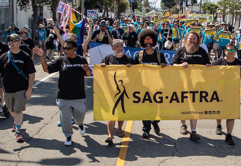 Miembros vistiendo camisetas negras "SAG-AFTRA" caminando en un desfile sosteniendo una pancarta amarilla SAG-AFTRA frente a ella.