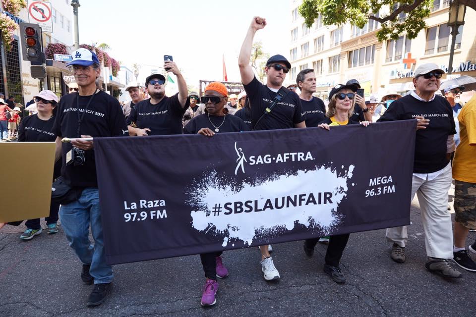 Miembros vistiendo camisetas negras de "May Day March 2017" marchando detrás de una pancarta negra que dice "La Raza 97.9 FM" a la izquierda, #SBSLAUNFAIR en negro sobre un diseño de salpicaduras de pintura blanca y "MEGA 96.3 FM" en blanco a la izquierda.