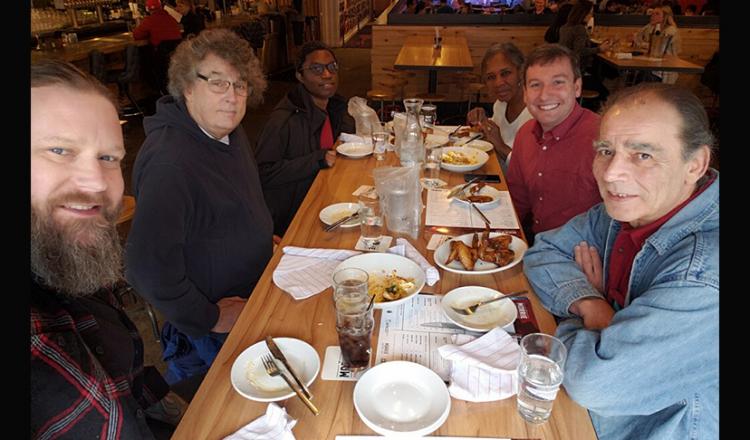 Un grupo diverso de hombres y mujeres se sienta a desayunar en un restaurante. Frente a la cámara, los miembros del grupo dan grandes sonrisas.