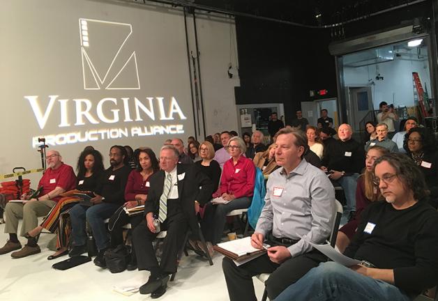 Un grupo de hombres y mujeres en los asientos mira a la izquierda fuera del escenario. Detrás de ellos, el logotipo de Virginia Production Alliance se proyecta en una pantalla.