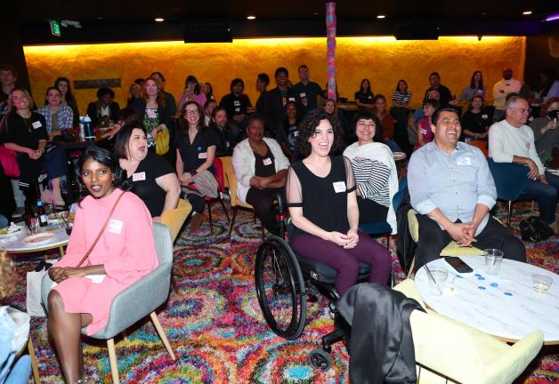 Un grupo diverso de hombres y mujeres se sienta en una audiencia. Una joven negra vestida de rosa, en el extremo izquierdo, está sentada a una mesa; otros están sentados en sillas o de pie. Todos miran hacia la extrema derecha.