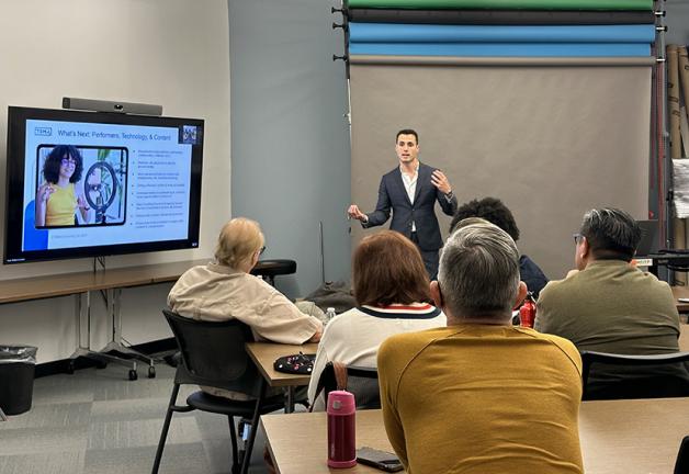 Un hombre con traje azul y camisa blanca enseña a un grupo de hombres y mujeres en una clase.
