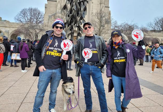 Los miembros del sindicato visten camisetas, chaquetas y gorras de SAG-AFTRA Strong, y sostienen carteles que dicen "¡Trabajadores graduados de BU en huelga!" Uno de los miembros también tiene un perro.
