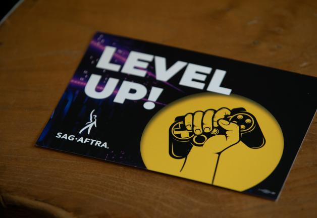 Una postal sobre una mesa. La postal muestra “¡Sube de nivel!” en letras blancas en mayúsculas, el logotipo de SAG-AFTRA y un gráfico de una mano alrededor de un controlador de juego.