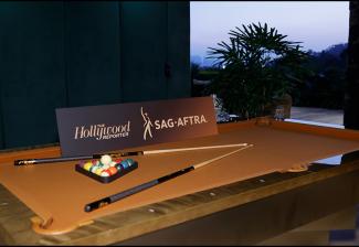 An atmospheric shot of a pool table. The pool balls and sticks are arranged stylistically. A placard with ‘The Hollywood Reporter’ and SAG-AFTRA logos is placed across the table. To the right is a palm tree against the setting skyline.