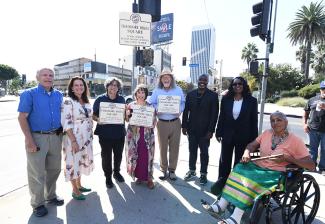 Zev Yaroslavsky, KatyYaroslavsky, Navarro, Ginsburg Bikel, Stein, Harris-Dawson, Wheeler and Calderon outside on the sidewalk. Navarro, Ginsburg Bikel and Stein hold up signs of the Theodore Bikel Square. Visible behind them on a street pole is the square’s street sign. 