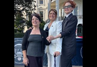 Bruno, Cyrille and Talkington outside on the White House lawn. Bruno wears a black cardigan over a gray top and black satin skirt; Cyrille wears a white pantsuit with a SAG-AFTRA enamel pin on the lapel and a black satin top; and Talkington wears a dark-colored suit. Bruno and Talkington hold hands in front of Cyrille, whose hand is on Bruno's shoulder. Behind the group is the front of the White House.