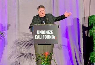 Astin se encuentra detrás de un podio con el cartel "Sindicalizar California". en letras blancas en mayúsculas en el escenario. Hace un gesto con el brazo izquierdo. El escenario tiene un telón y algunas plantas y vegetación.