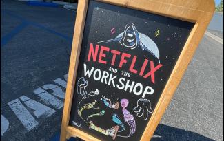 A photo of an outdoor blackboard-style sign in a parking lot. The hand-designed sign reads “Netflix and the Workshop” in all caps, with “Netflix” in red. There is also a cartoon sketch of a boy falling into bed as various characters watch his descent. 