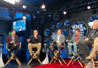 Crabtree-Ireland, seated second from left, speaks. He wears a black jacket with SAG-AFTRA’s logo and khaki pants. Other panelists are visible. The studio has bright lights, KNX’s logo, and a red carpet on the floor.