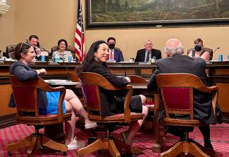 Long (asiático-americana) se sienta en una silla mientras sonríe por encima del hombro. Al fondo, los senadores están sentados detrás de un estrado.