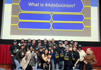 Una foto de un grupo grande y diverso en el escenario debajo de una pantalla de cine. Muchos visten camisetas solidarias negras de SAG-AFTRA. Sobre ellos, en la pantalla de cine, está la copia: "¿Qué es #AdsGoUnion?" y cuatro espacios en blanco debajo.