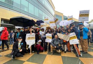 A large group of members outside in the rain. Each wears protective or winter clothing to stave against the rain; a few show their SAG-AFTRA strike apparel. Picket signs and umbrellas seen throughout. 