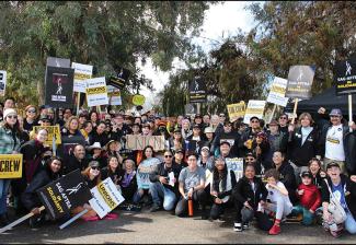A very large, diverse group outside in a park setting. Those in the front two rows either sit or kneel on the concrete. Visible throughout are black and white SAG-AFTRA picket signs; there is also a cardboard sign with "Union" in large, all-cap copy. 