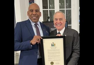 Knight (Black) and DiNapoli hold up a framed certificate. Knight is in a blue blazer and light blue dress shirt; DiNapoli is in a dark-brown blazer, a white dress shirt and tie. 
