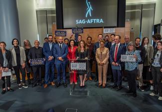 A large group of people stand in an auditorium-like room. Many hold signs. Behind the group is a projector screen bearing the SAG-AFTRA logo in white.