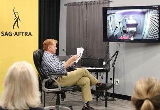 A man with red hair is seated in a black office chair at a desk, reviewing a script. He is wearing a blue-and-white checkered shirt, khaki pants and black shoes with colorful patterned socks. A microphone and various pieces of audio equipment are set up on the desk, and a laptop is open in front of him. To the left, a yellow SAG-AFTRA banner with its logo is displayed.