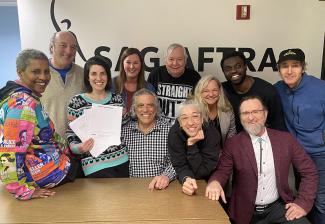 A group of people gather around a table, in front of a wall painted with the SAG-AFTRA logo.