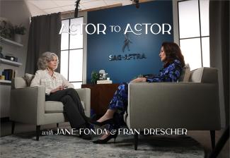 Jane Fonda and Fran Drescher are seated in a room and captured mid-interview. The two face each other and can be seen speaking with one another.