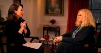 Drescher and Streisand, seated in a decadent sitting room. Both women face each other. Drescher's hands are clasped in front of her and she smiles at Streisand. A sheet of paper rests on her lap.