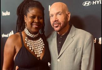 Two individuals are on the red carpet. The person on the left wears a dress and a cowrie shell necklace; the person on the right wears a gray blazer and a black dress shirt. Behind them is a black step-and-repeat backdrop.