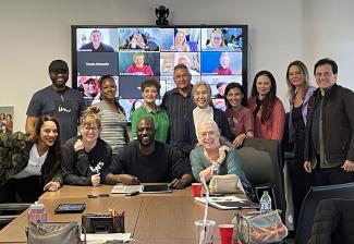 Attendees pose for a photo in front of a screen showing those who are attendning via Zoom. Some members sit at a table that has various items on it.