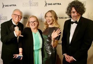 Dos hombres con esmoquin y dos mujeres con vestidos posan frente a un telón de fondo con logotipos de SAG-AFTRA.