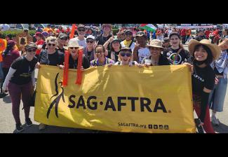 A large group of people of different ethnicities pose with a yellow SAG-AFTRA banner outside. Many wear SAG-AFTRA or "SAG-AFTRA Strong" T-shirts.