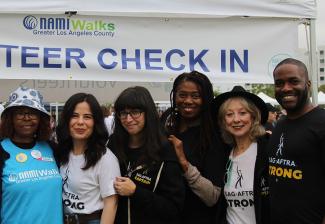 Seven people of different ethnicities outside of a tent. Aside from a Black woman, second from left, who wears a sky blue NAMIWalks T-shirt, all wear black or white “SAG-AFTRA” T-shirts. The tent banner reads “Volunteer Check In” in all caps and shows the