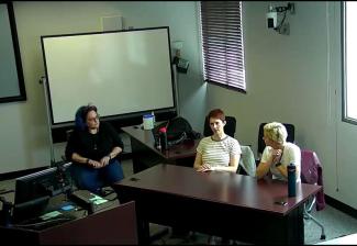 Three individuals are in a classroom setting. Two sit behind a table while one on the far left is in an office chair. The person on the far right is captured mid-speech while the other two listen intently.