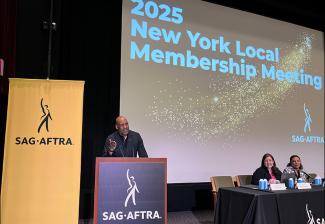 Knight stands on a podium with SAG-AFTRA on a black background in white text. To his left is a yellow banner with SAG-AFTRA in black text. There is a large screen behind him with the “2025 New York Local Membership Meeting” title card. Two individuals sit to his right behind a table. 