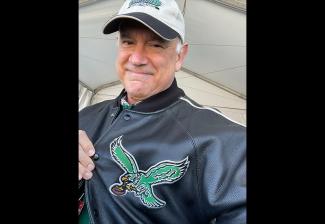 Una selfie de Macnow. Es un hombre mayor con cabello blanco. Lleva una gorra deportiva blanca y una chaqueta tipo letra negra. Se tira de la solapa de su chaqueta para mostrar la imagen de un águila verde con las alas extendidas sosteniendo una pelota de fútbol entre sus garras.