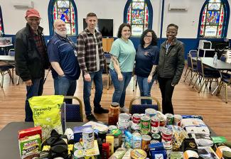 Six people, three men and three women, stand in a church behind a table piled with food. There are three stained glass windows in the background.