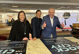 Three people smile in front of a marble table that has two black and blue posters that read “Rock the City.” The person on the right is giving thumbs up. 