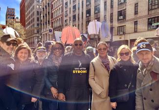 The group, wearing warm-weather jackets, pose for a picture on the street. Shirts on poles can be seen in the background.