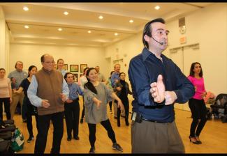 A gentleman on the right pantomines a dance move. Behind him, a diverse group of men and women, follow his movements..