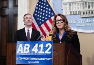 Fisher is behind a podium with a sign for AB 412. She wears a blazer over a blue shirt, jewelry and glasses. She holds the microphone’s neck as she speaks. Behind her, a person listens intently. Also visible is the American flag and an image of the California State Capitol building.
