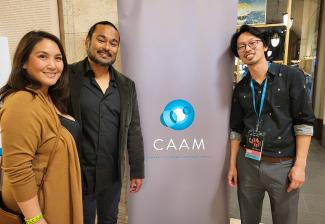 From left, Ocampo-Shah, Shah and Hsieh in a lobby-like setting. They frame a standing banner for the Center of Asian American Media, which shows its logo, an abstract drawing of interconnected circles in shades of blue and white, and copy “CAAM” in white.