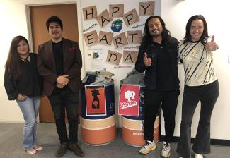Two men and two women stand next to the two collection bins. On the wall behind them are letters that spell out "Happy Earth Day." 