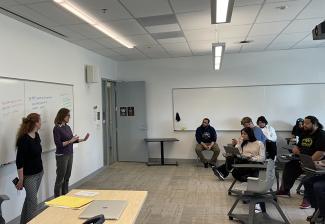 Escobedo, left, and Hausherr speak to college students in a classroom. Both women are in front of a whiteboard on the far left; the students are in desks on the far right. As Hausherr speaks, she gestures with her hands. 