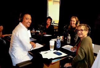 David white sitting at a table with headphones wearing a white button up smiling sitting next to Carteris in a olive Cardigan and headset. Bender and Harcharic sit side by side and across the table from White with headphones on.