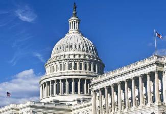 An image of the United States Capitol from a side profile. Background: blue skies with soft white clouds scattered throughout the sky.