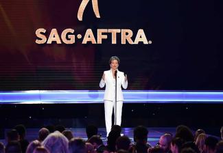 Carteris in white suite on a stage speaking into a mic looking out at the audience