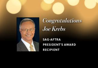 Joe Krebs headshot. Krebs in white button up, tan tie and dark brown coat in front of a dark background. 