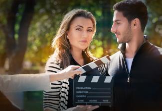 Image of a female looking at the camera and a gentleman looking at her with a nature background. In the foreground is a person holding a clapper.