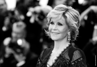 A black and white photo of Jane Fonda on the red carpet. She is positioned on the left of the image and turned towards the right. Behind her are blurred images of photographers.